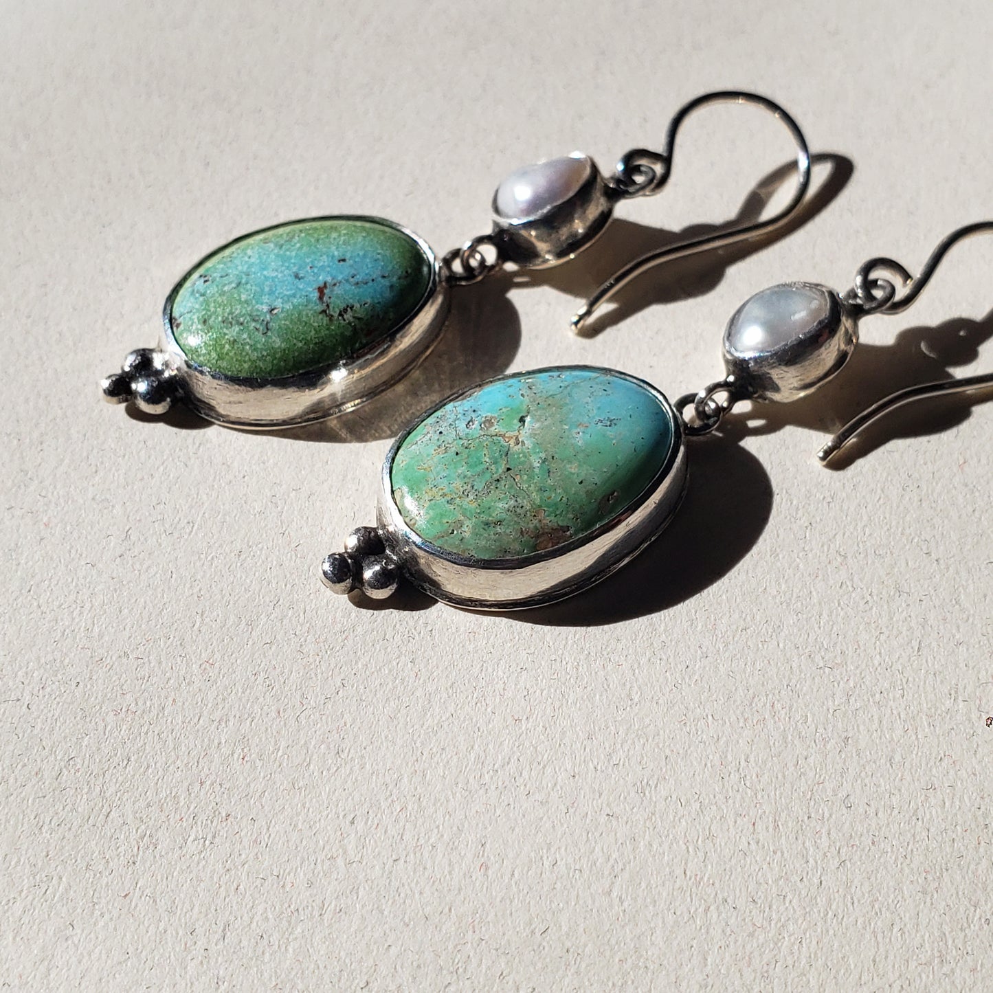 This is an image of a pair of oblong turquoise earrings with pearls. The top portion is a Keshi pearl and the bottom is oblong, oval Rosa Verde turquoise. These earrings are lightweight and have beautiful muted tones that emit a calm feel. They measure 2" in length x .50" width and are handcrafted from sterling and fine silvers by Jennifer Favour.
