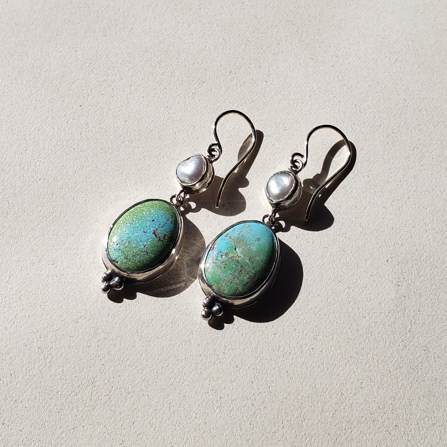 This is an image of a pair of oblong turquoise earrings with pearls. The top portion is a Keshi pearl and the bottom is oblong, oval Rosa Verde turquoise. These earrings are lightweight and have beautiful muted tones that emit a calm feel. They measure 2" in length x .50" width and are handcrafted from sterling and fine silvers by Jennifer Favour.