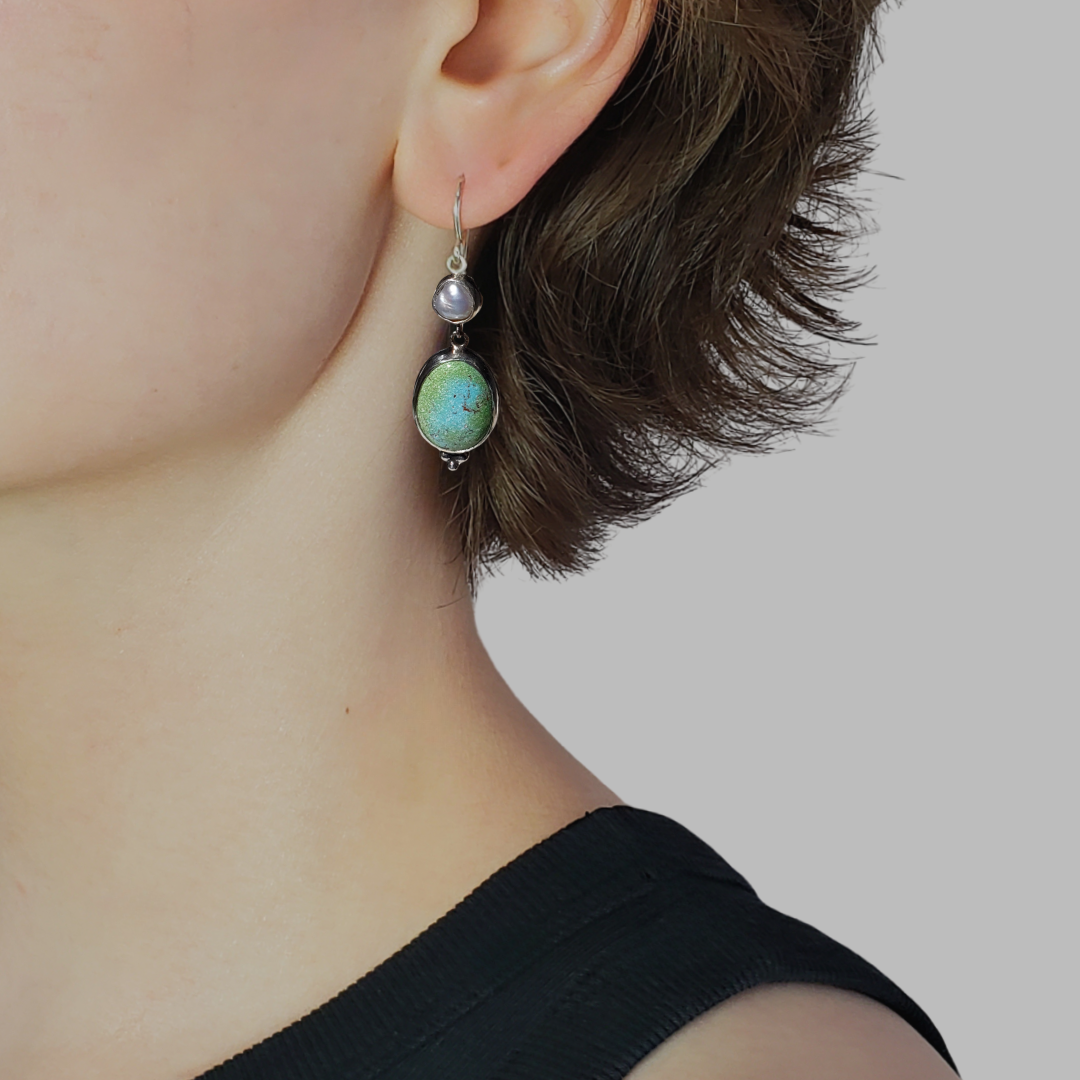 This is an image of a model wearing a pair of oblong turquoise earrings with pearls. The top portion of these earrings is a Keshi pearl and the bottom portion is an oblong turquoise Rosa Verde gemstone. These earrings are lightweight and have beautiful muted tones that emit a calm feel. They measure 2" in length x .50" width and are handcrafted from sterling and fine silvers by Jennifer Favour.