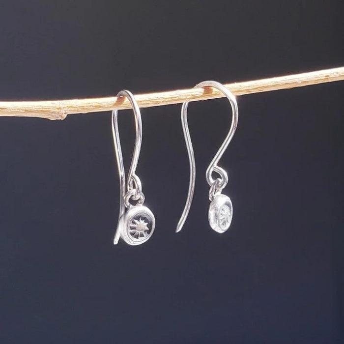 A right facing silhouette of a pair of small sterling silver artisan earrings hung from a bamboo stick against a dark gray background.