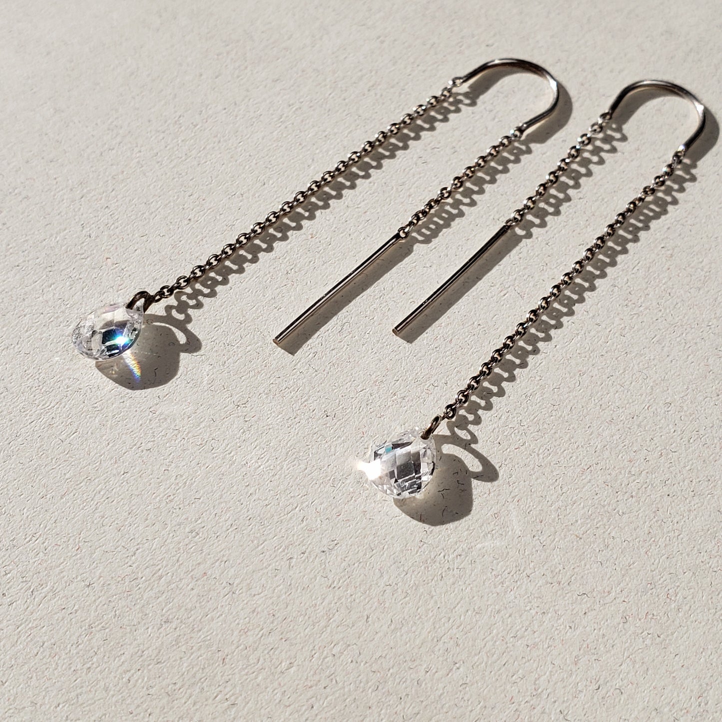 Long silver threader earrings with a cubic zirconia teardrop briolette drop laying on a flat surface left facing.