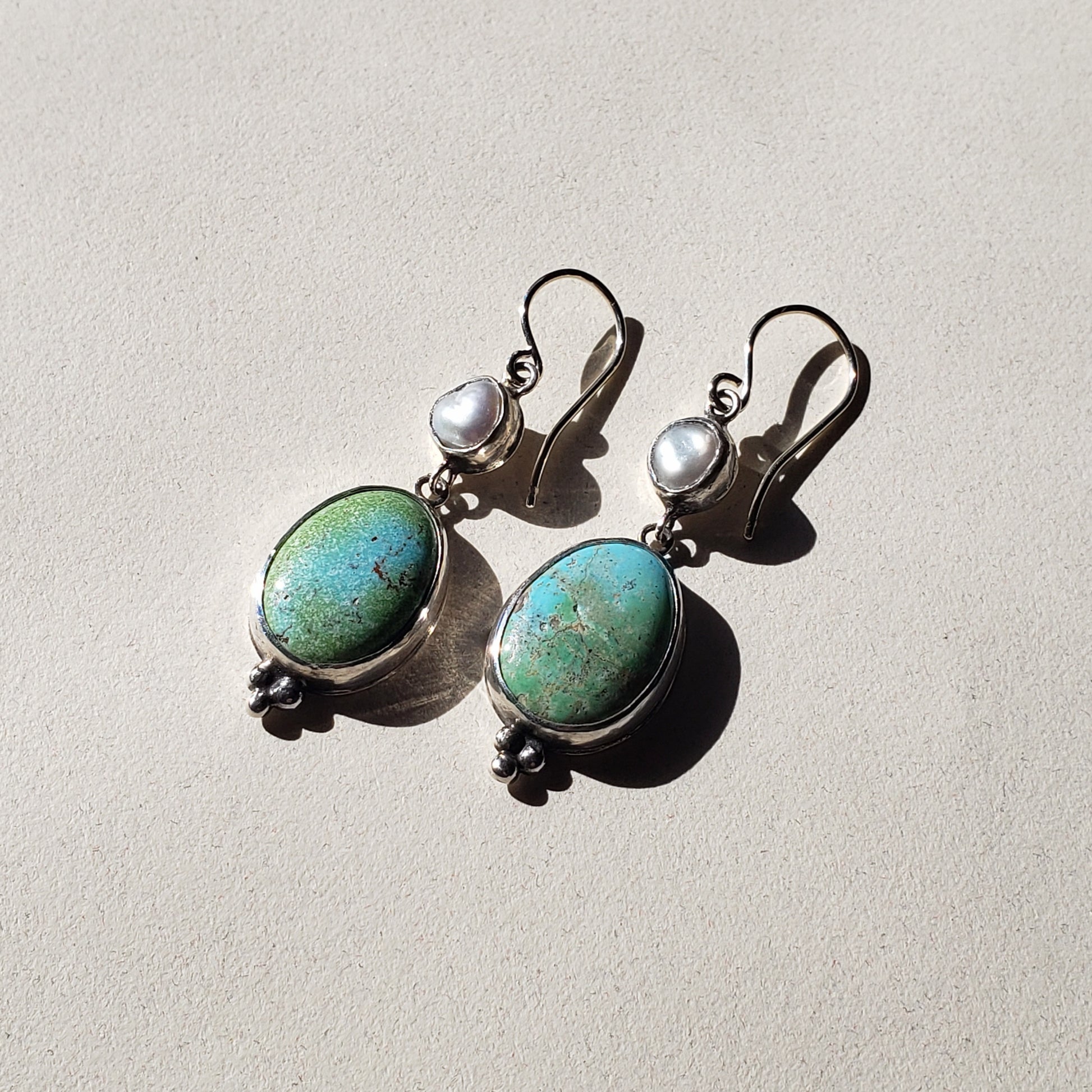 This is an image of a pair of oblong turquoise earrings with pearls. The top portion is a Keshi pearl and the bottom is oblong, oval Rosa Verde turquoise. These earrings are lightweight and have beautiful muted tones that emit a calm feel. They measure 2" in length x .50" width and are handcrafted from sterling and fine silvers by Jennifer Favour. 