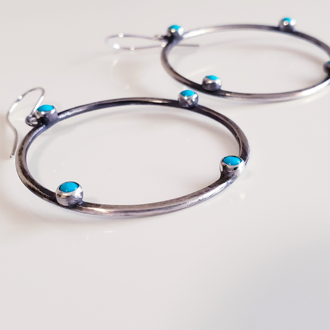 Image of sterling silver hoop earrings with small turquoise gemstones. Hoop earrings are laying on white board for display. 