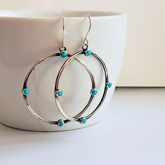 Image of sterling silver hoop earrings with small turquoise gemstones. Hoop earrings are laying on white board for display. 