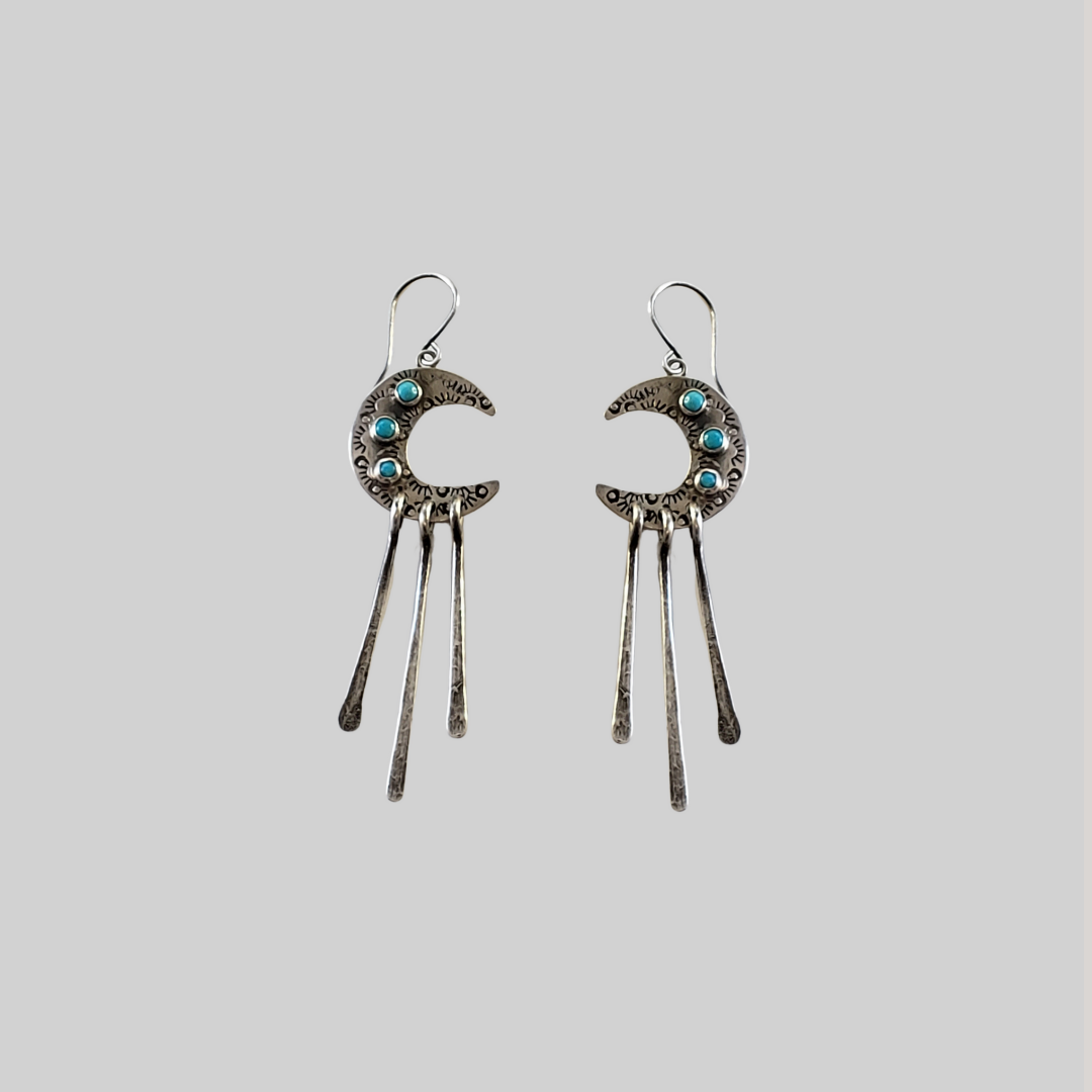 Silver crescent moon earrings with turquoise stones and three long dangles laying on gray surface 