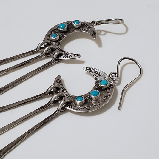 Silver crescent moon earrings with turquoise stones and three long dangles laying on gray surface