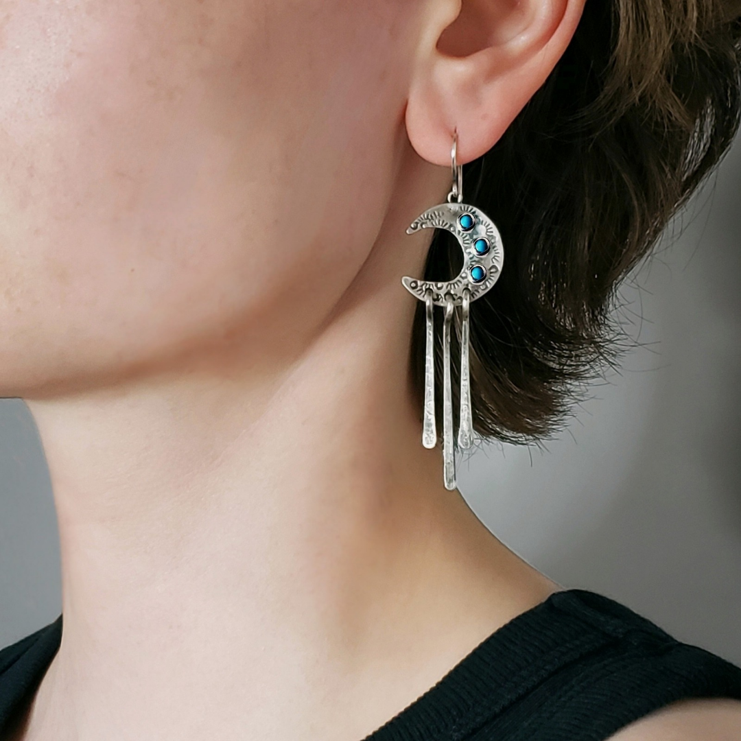 Left facing woman wearing silver crescent moon earrings with turquoise stones and three long dangles laying on gray surface