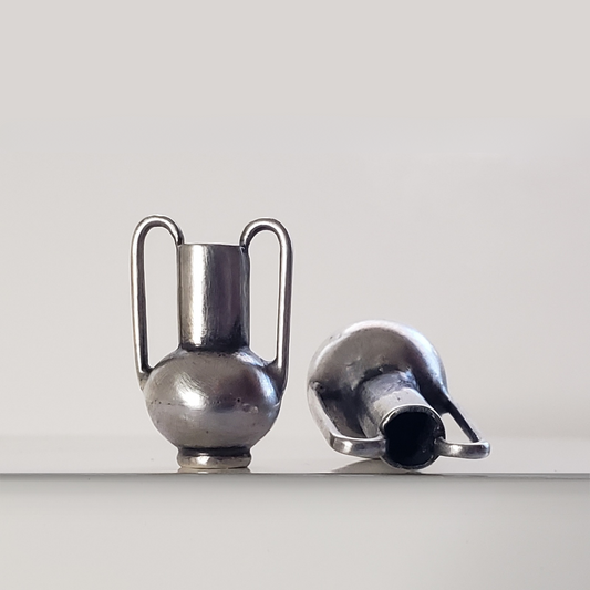 Image of two handmade sterling silver vessel pendants sitting on white display. Vessel on the right is tipped on its side. 