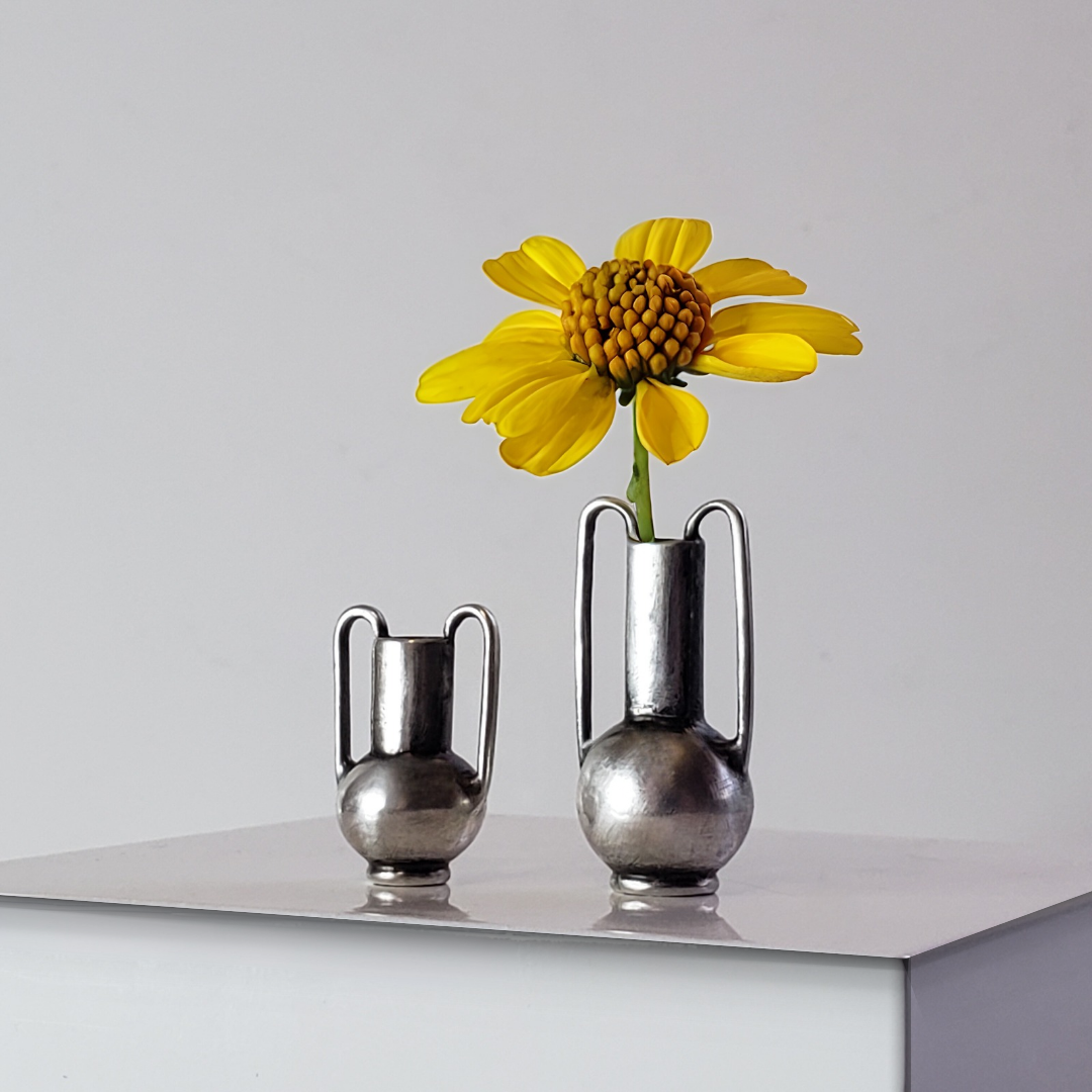 Image of two handmade sterling silver vessel pendants sitting on white display. Vessel on the right has a yellow daisy inside of jt. 