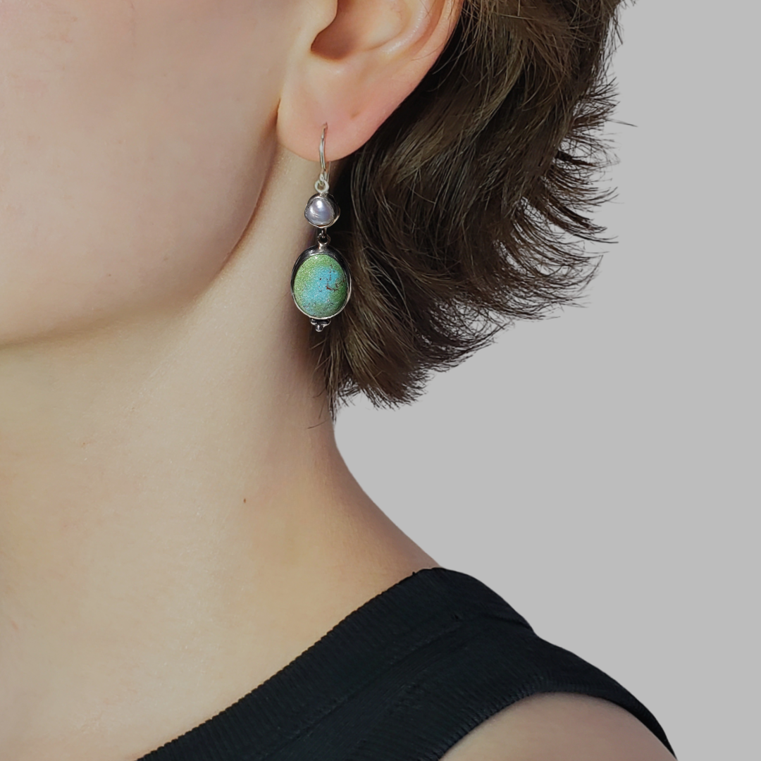 This is an image of a model wearing a pair of oblong turquoise earrings with pearls. The top portion of these earrings is a Keshi pearl and the bottom portion is an oblong turquoise Rosa Verde gemstone. These earrings are lightweight and have beautiful muted tones that emit a calm feel. They measure 2" in length x .50" width and are handcrafted from sterling and fine silvers by Jennifer Favour. 