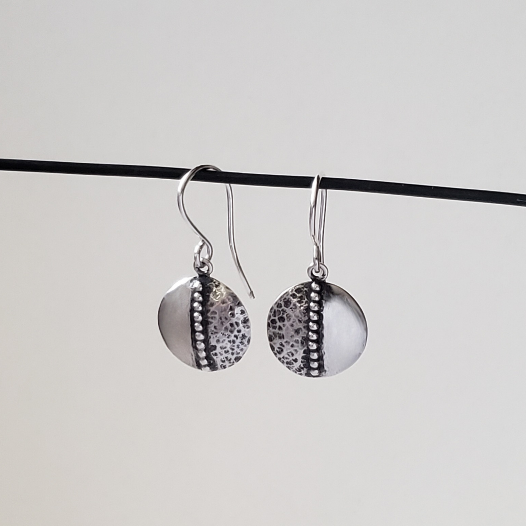 This is an image of a pair of small textured silver disc earrings. These silver disc earrings feature a half texture design, while the other half of the disc remains smooth silver. Down the center of the disc earring is a beaded accent piece, dividing the circle into two parts. These are all handcrafted silver earrings and are a featherweight style, light and easy to wear. They measure approximately half an inch in diameter and are hung from sterling wires. All handmade earrings, all sterling silver. 