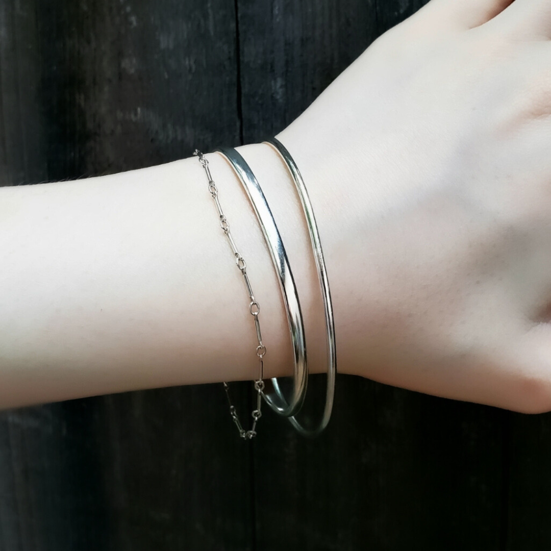 Womans wrist with a shiny silver stacker bracelet and two dainty silver bracelets against a black wooden fence 