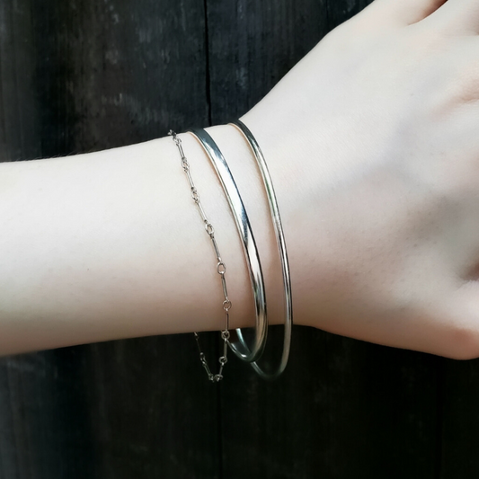 Womans wrist with a shiny silver stacker bracelet and two dainty silver bracelets against a black wooden fence 