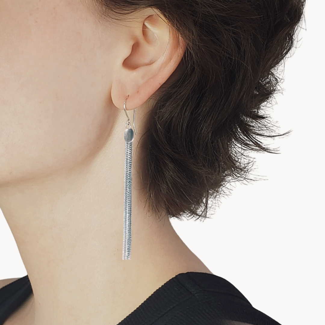 Left facing woman wearing long silver tassle earrings against beige background 