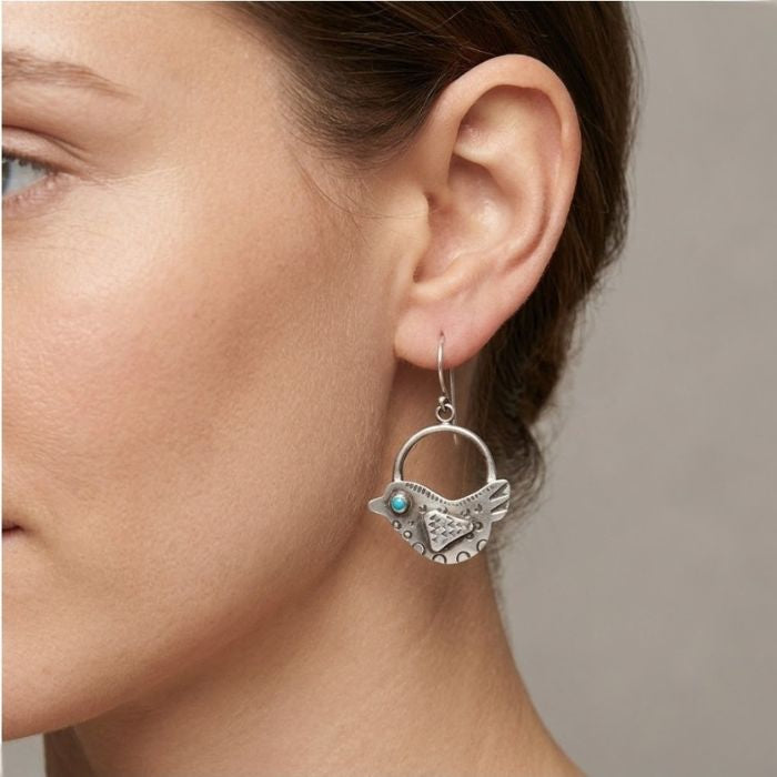Close-up of a woman wearing a silver bird-shaped earring with a turquoise stone on a neutral background.